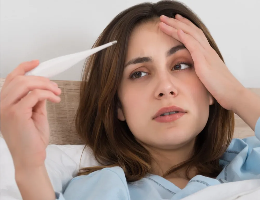 Person holding a thermometer and touching their forehead, indicating fever and discomfort, common in Rheumatoid Arthritis (RA)